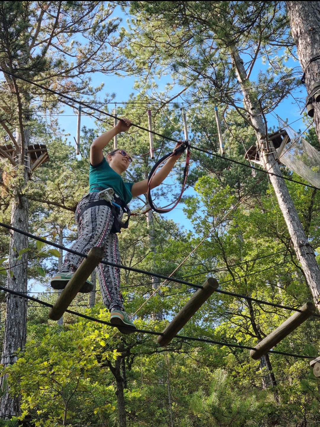 Une jeune Aspirante Chevalier 🛡️ progresse avec concentration sur le pont de rondins espacés du parcours marron "L'Équilibre du Baron". Au-dessus d'elle, on aperçoit les obstacles suspendus du parcours noir, créant une impression de hauteur et de défi entre les différents niveaux du parc. accrobranche Nyons