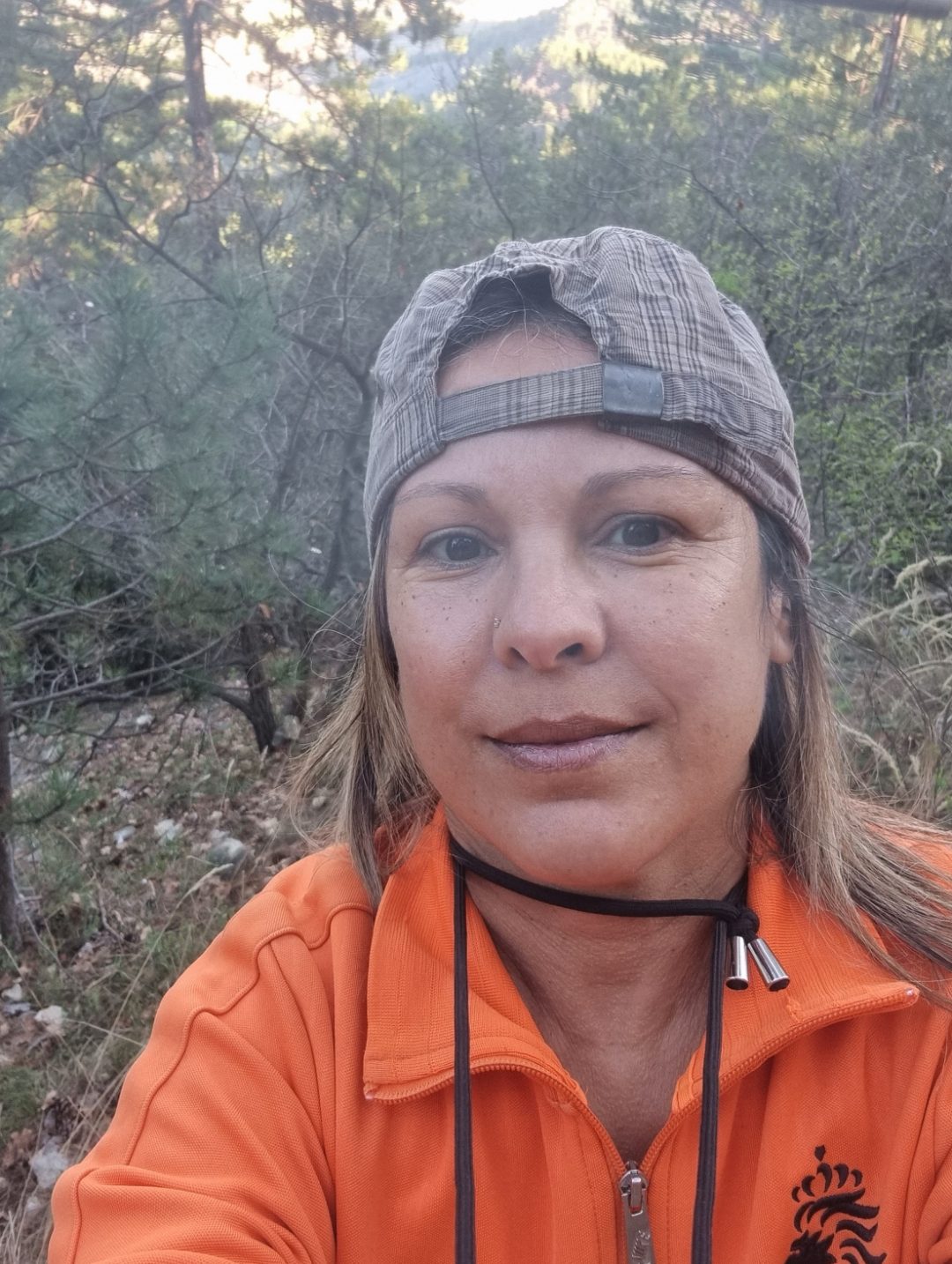 Portrait simple et authentique de la Baronne 👸, responsable du parc accrobranche à Nyons 🌲. Elle se tient debout sur un parcours en bois, portant son équipement de sécurité, et regarde avec un sourire chaleureux un participant. L'arrière-plan est la forêt de pins, sans vue dégagée.